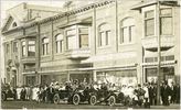 Original title:  Courtesy Saskatoon Public Library. Crowd in front of J. F. Cairns store on 2nd Avenue South admire passing automobiles. between 1906 and 1912
